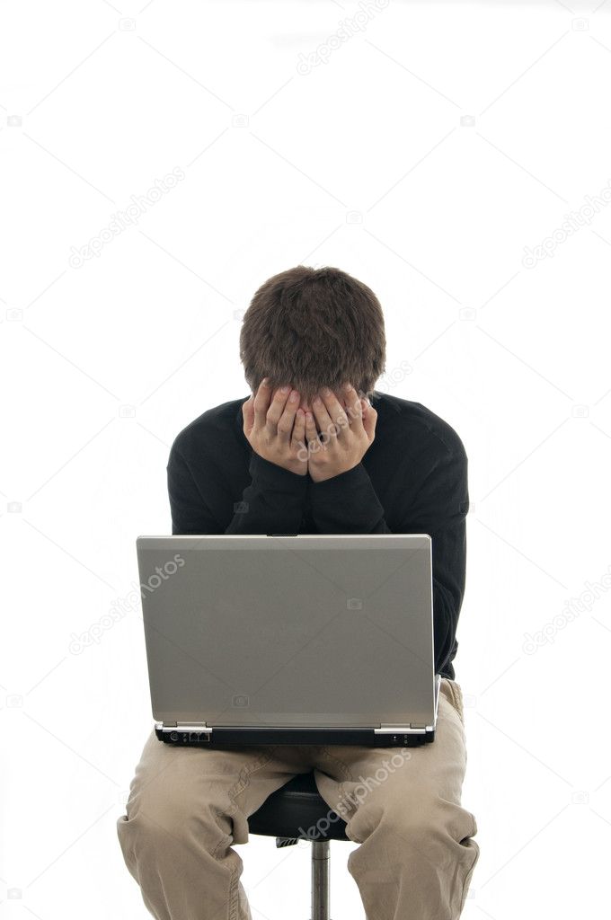 Teenager sitting on stool with laptop and his hands covering his face on white background