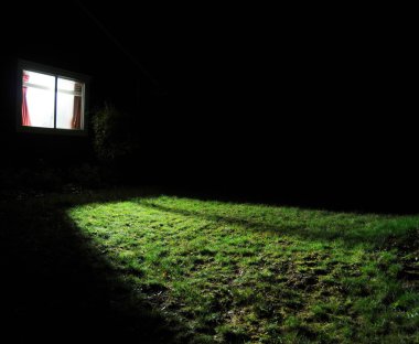 A dark house at night with a window with a light on in the room spilling out light onto the grass lawn yard. clipart