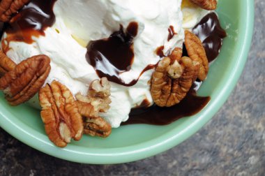 A sundae with pecans, hot chocolate fudge, whip cream, and ice cream inside of a green dessert bowl sitting on a kitchen counter top. clipart