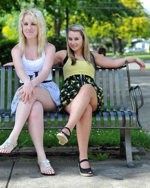两个女孩朋友坐在金属的长凳上 在对照相机微笑 他们可以被视为轻浮 快乐或欢迎 — 图库照片