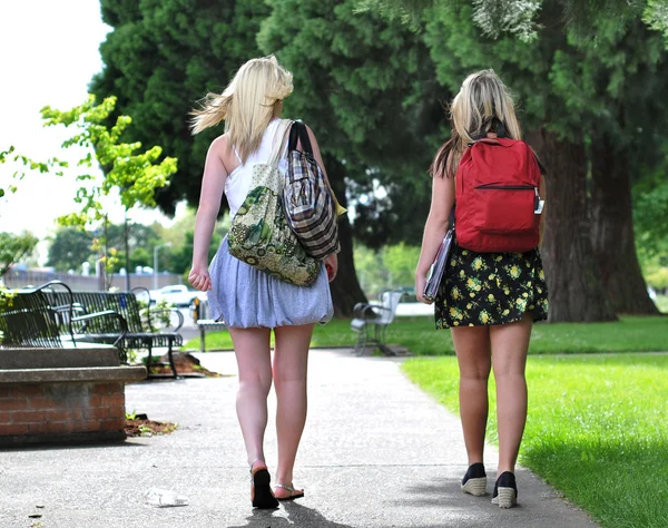 两个年轻有魅力女孩穿裙子走在前面一所公立学校与他们背的包上他们 — 图库照片