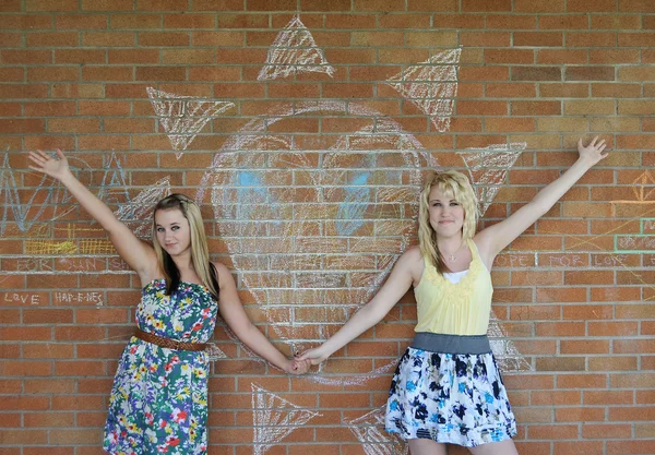 两个美丽白色白种人金发碧眼的女孩站在砖的墙上用粉笔写的心设计旁边 — 图库照片