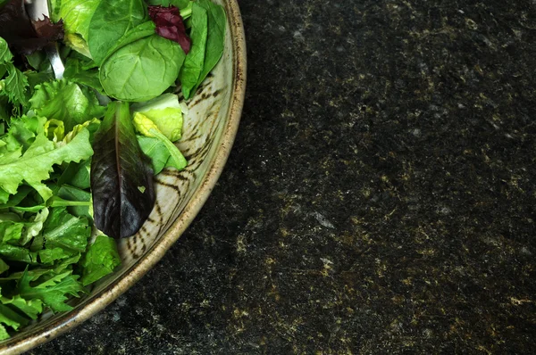 stock image A variety of green lettuce tossed in a salad bowl, sitting on a black kitchen counter top. Copyspace on the right.