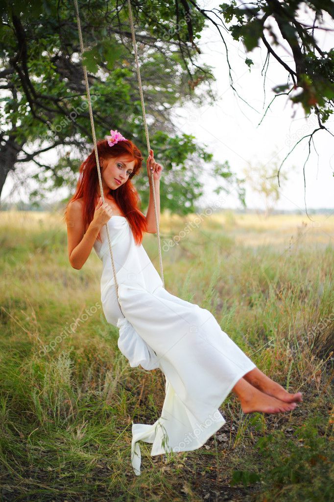 The red-haired girl in a white dress shakes on a swing in a grove