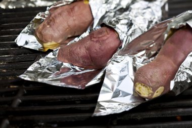 Cooking sweet potatoes wrapped in tin foil over a gas grill. clipart