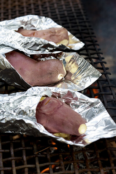 Sweet potatoes wrapped in aluminum foil are being cooked over an open fire.