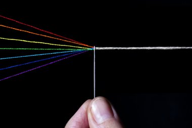 Threads of cotton in the colour spectra going through the eye of a needle with finger holding the needle, turing from colours to white thread clipart