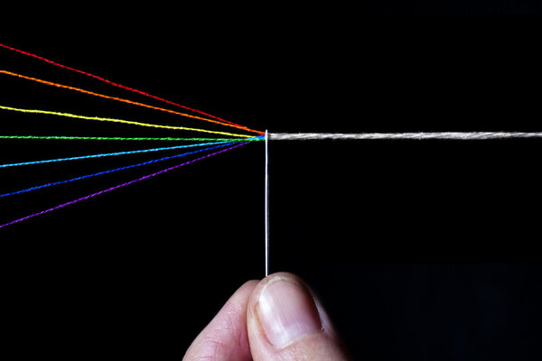 Threads of cotton in the colour spectra going through the eye of a needle with finger holding the needle, turing from colours to white thread