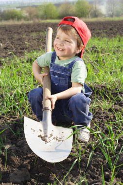 Smiling little boy sitting on field with shovel clipart