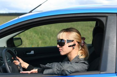 Young blond woman in a blue car in sun-glasses with hands free bluetooth clipart