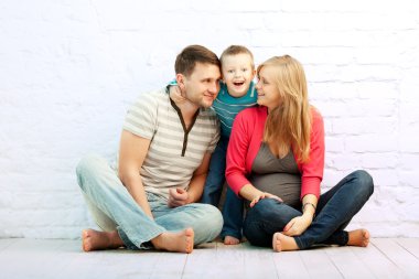 Happy family of three sitting on the floor near the wall: mother, father and little boy. Mother is pregnant. Son is wondered. clipart