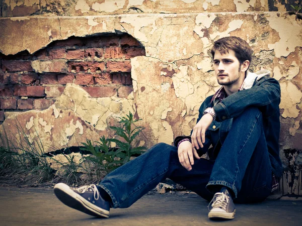穿牛仔裤的衣服的年轻男子坐在裂损毁墙前地面上 免版税图库图片