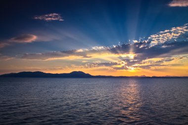 Midterran Denizi 'nin üzerinde turuncu gün batımı, yukarıdaki derin mavi gökyüzü, koyu yoğun bulutların arasında çimdiklenen güneş ışınları, deniz dalgalarının yansıması, Arnavutluk kıyıları