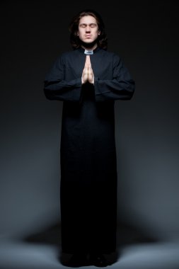 Young priest is praying against dark background clipart